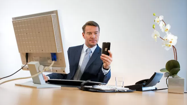 Hombre frente al computador, distraído con el celular en la mano, mientras trabaja en su oficina