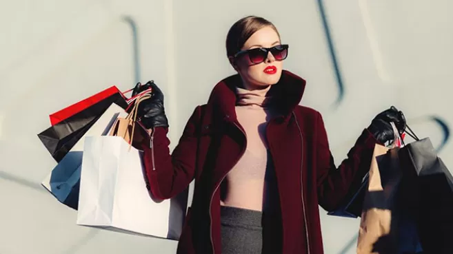 Mujer elegante con saco color vino tinto y gafas oscuras, cargando varias bolsas de compras en ambas manos