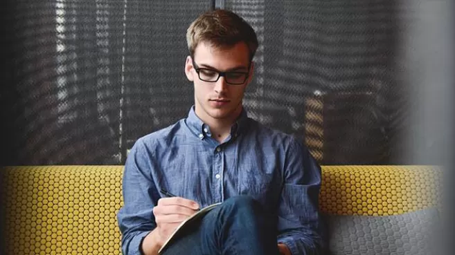 Hombre joven con anteojos, vestido de azul, sentado escribiendo