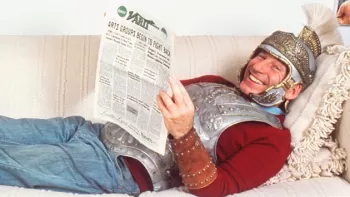 Mel Brooks sonriendo vestido con armadura de Caballero, acostado en un sofá sosteniendo un periódico abierto