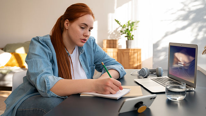 Mujer joven escribiendo en una libreta mientras estudia virtualmente desde su laptop