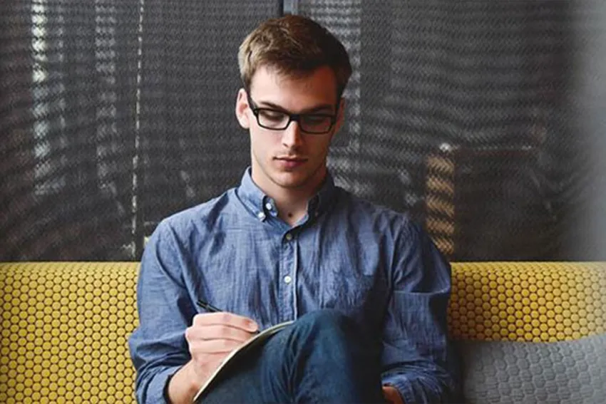 Hombre joven con anteojos, vestido de azul, sentado escribiendo