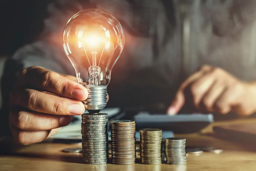 Manos de hombre sosteniendo una bombilla encendida sobre pilas de monedas