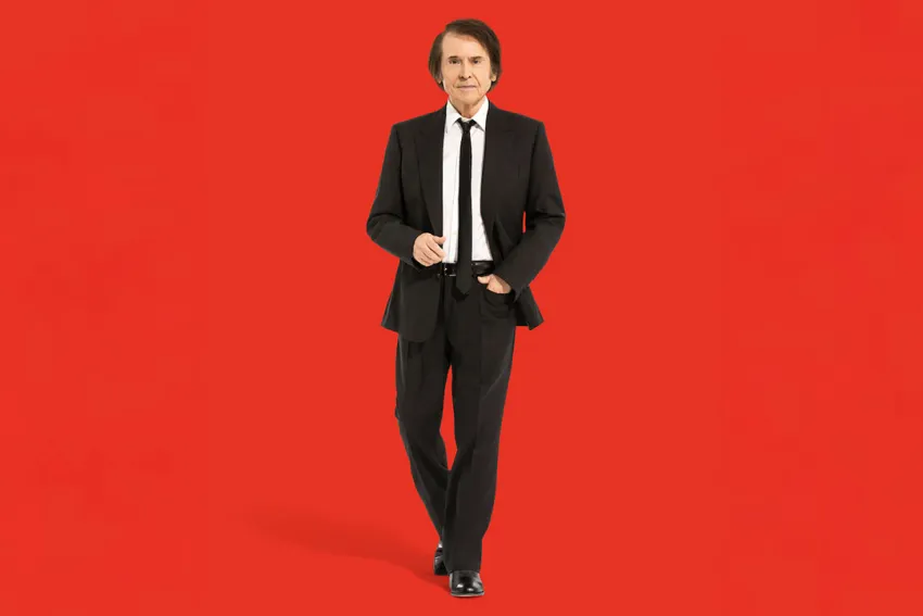 Raphael vestido de traje negro, y de fondo una pared roja