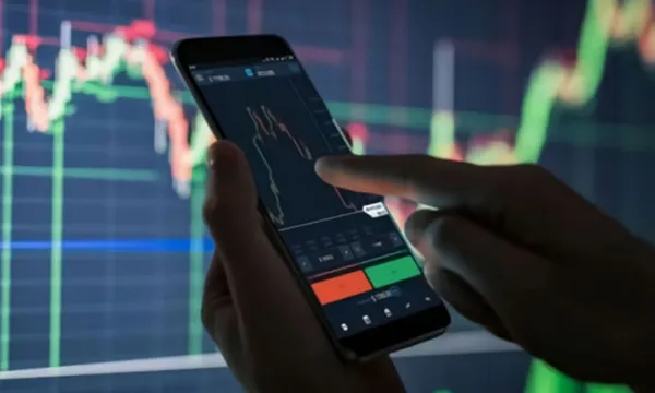 Manos haciendo trading desde el móvil y de fondo el gráfico del mercado