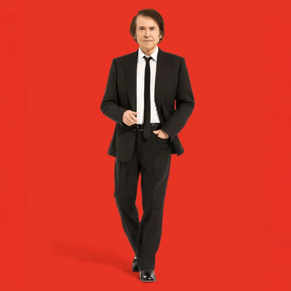 Raphael vestido de traje negro, y de fondo una pared roja